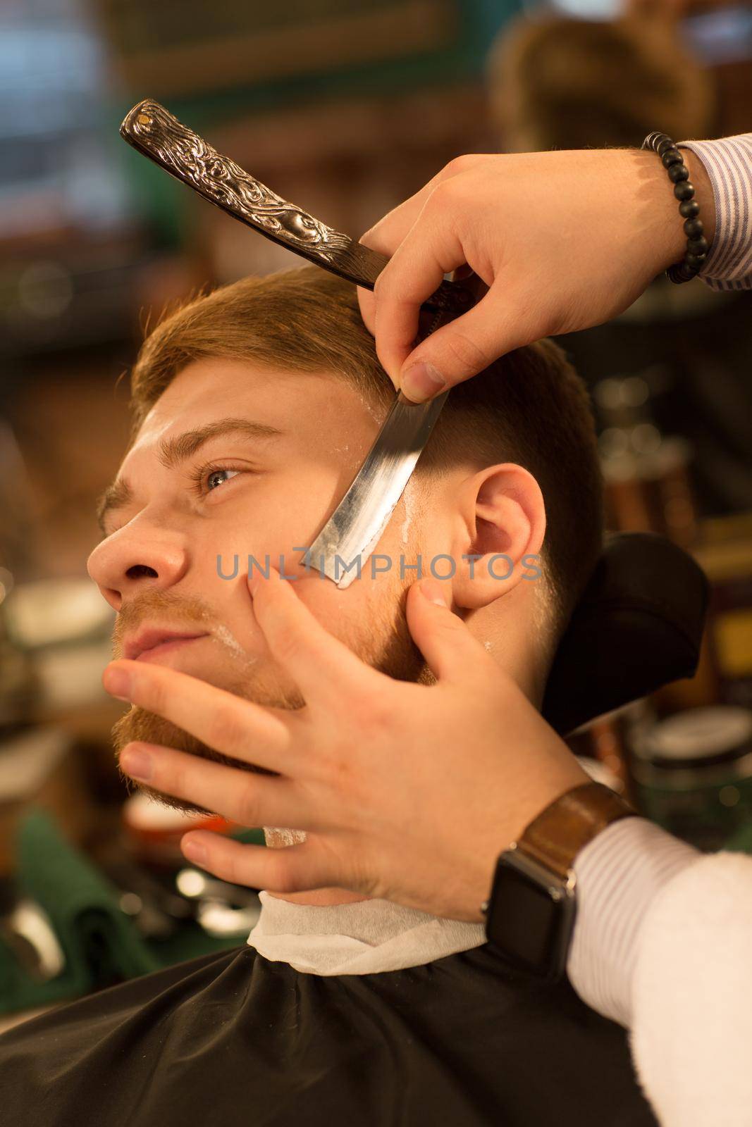 Vertical cropped close up of a young bearded man getting his beard shaved by a professional barber using razor barbering barbershop service lifestyle hipster classic traditional retro blade vintage.