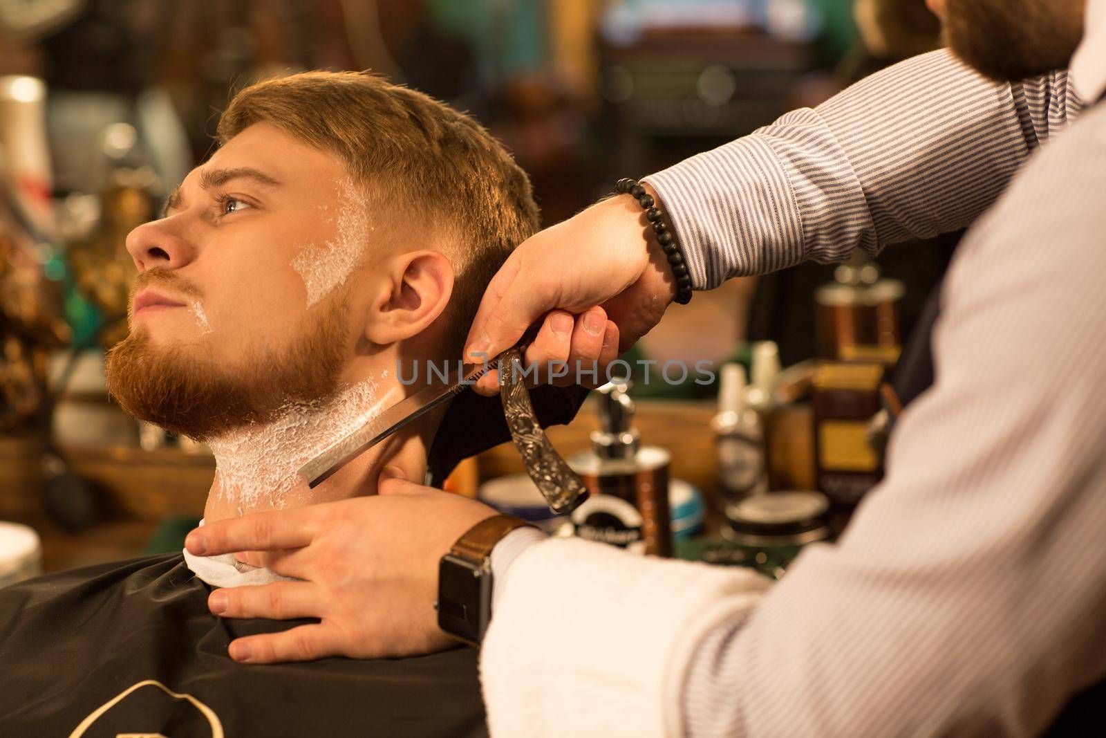 Close up of a young handsome bearded man looking away having his beard shaved by a professional barber with a razor service barbering profession occupation lifestyle hipster classic vintage retro wellness.