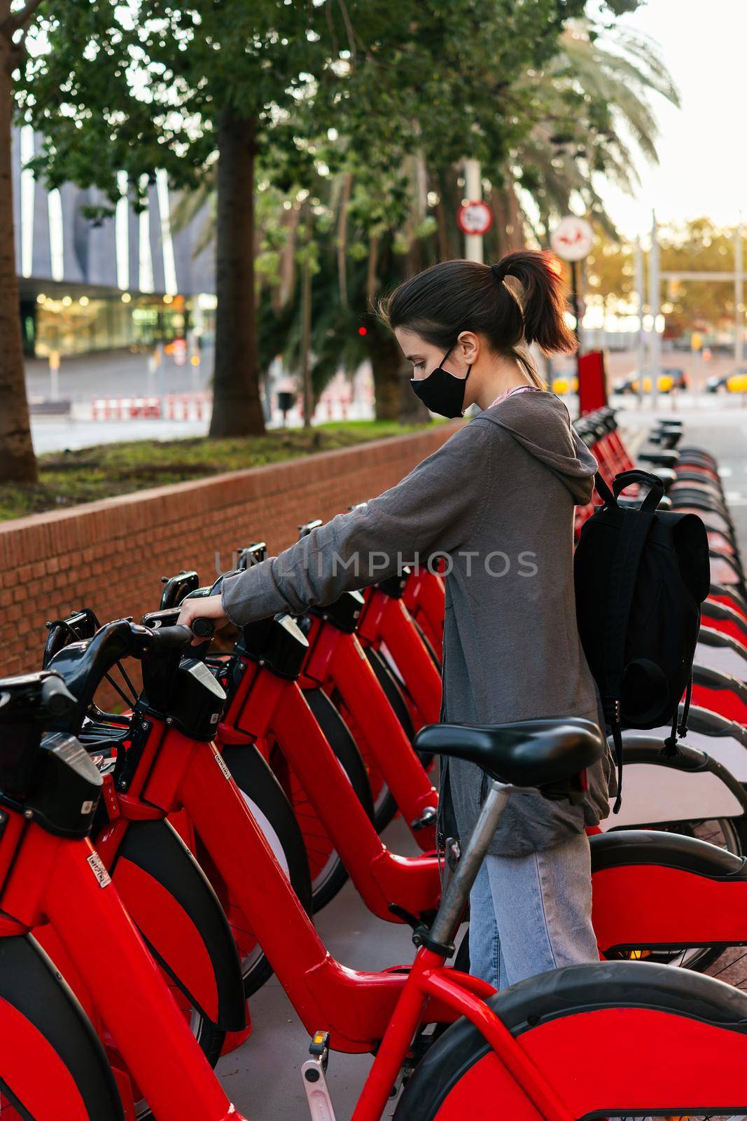 vertical photo of a young girl with protective mask taking a bike from an urban bicycle rental parking, concept of active lifestyle and sustainable mobility