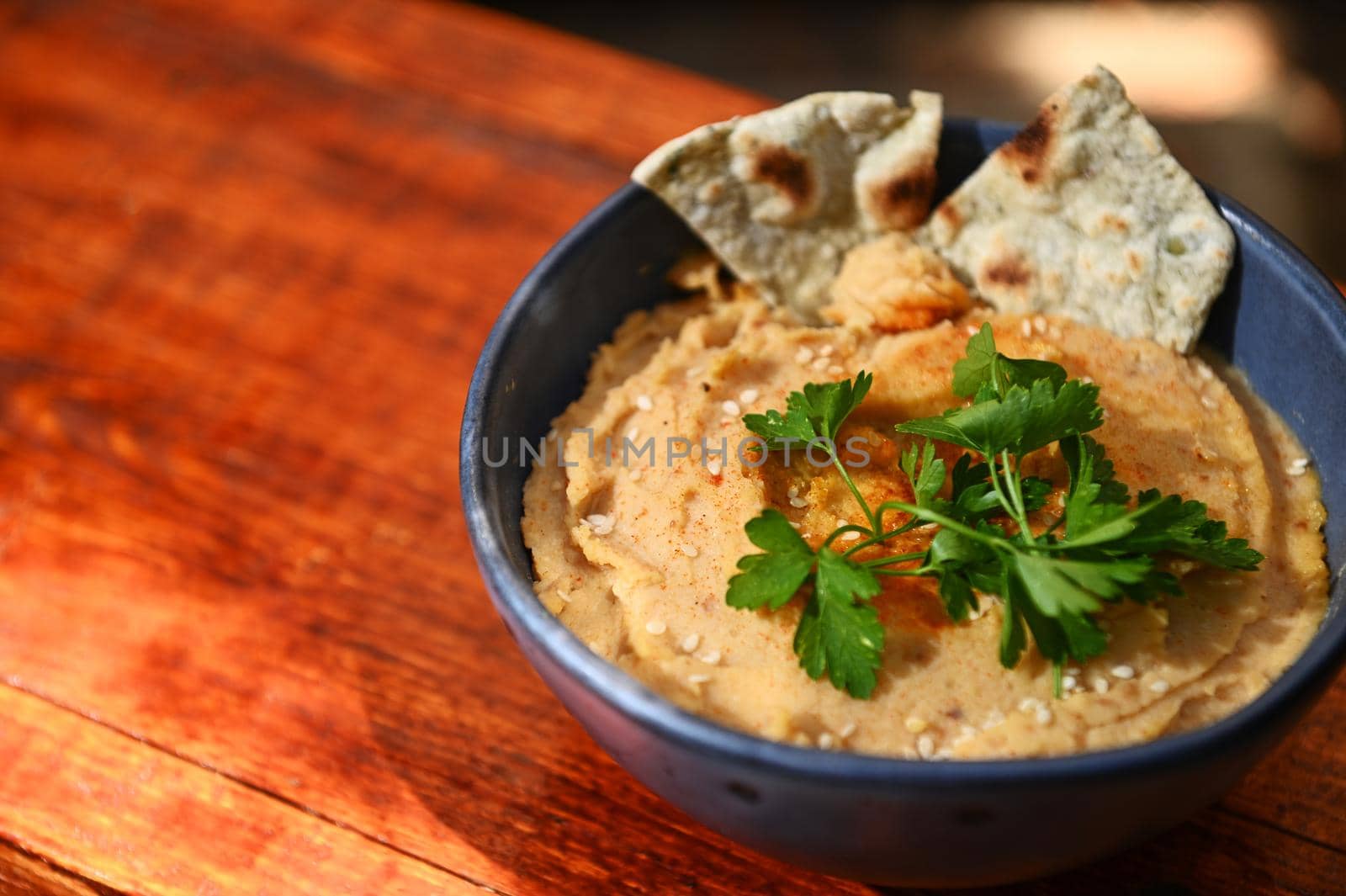 Close-up of a chickpea hummus dip with pita bread, parsley and sprinkled paprika on a blue ceramic bowl, on a rustic wooden table background with copy ad space for text. Still life. Food composition