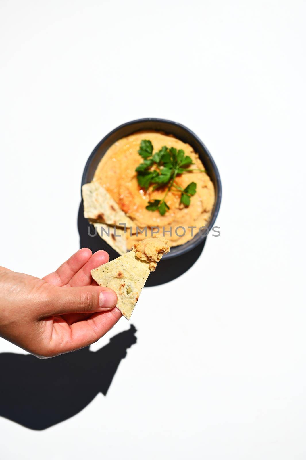 Overhead view. Vertical studio shot of a hand, dipping pita bread into a creamy consistency vegan dish - oriental hummus with parsley in blue ceramic bowl, isolated on white background.