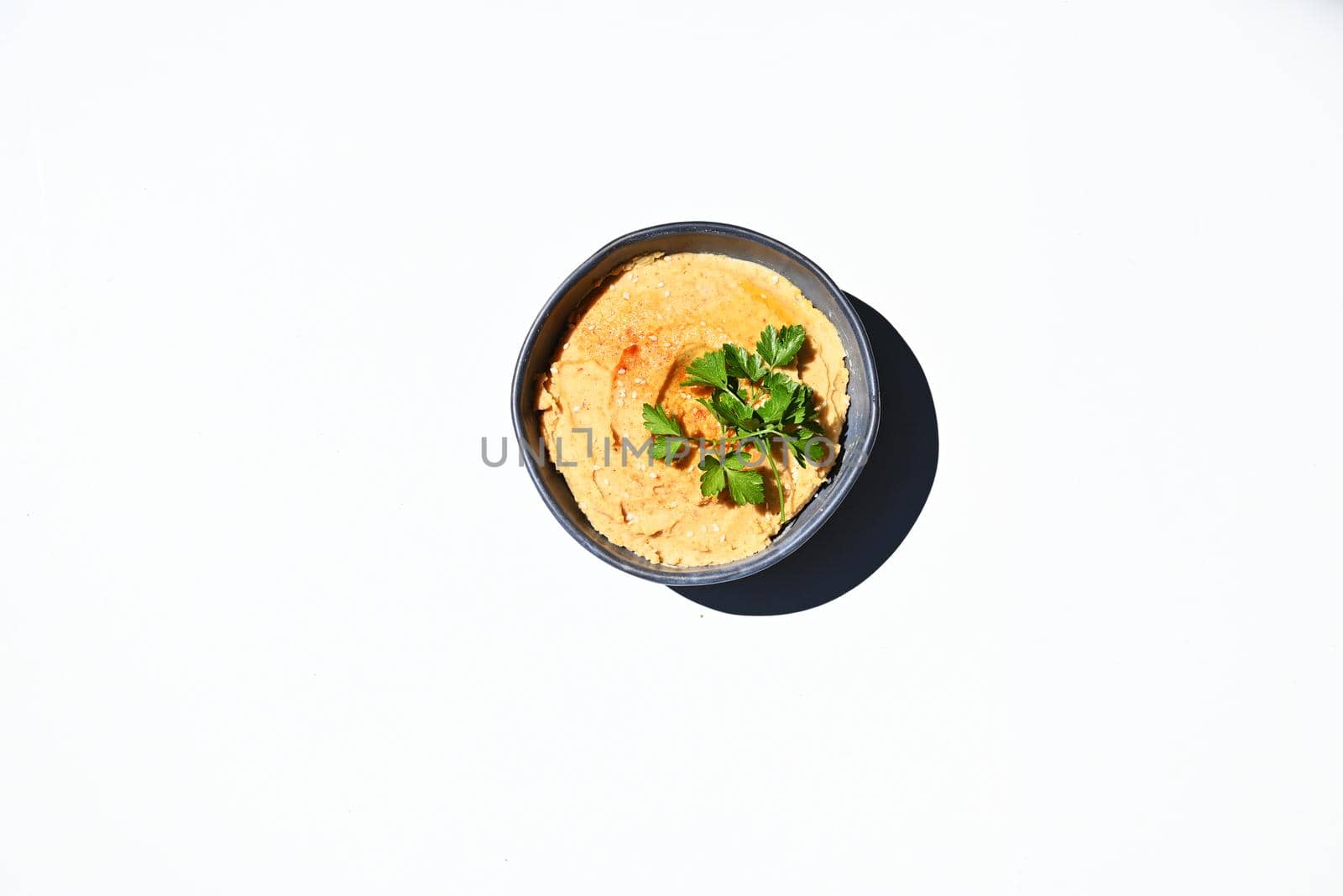Still life. Flat lay composition of a blue ceramic bowl of hummus sprinkled with paprika, parsley and olive oil, isolated over white background with copy space for advertising text