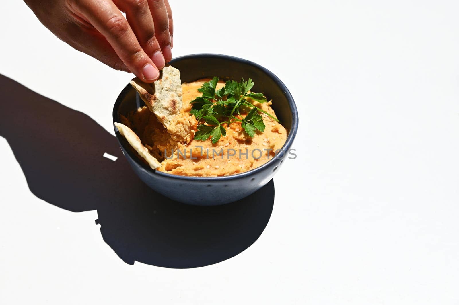 Close-up. Horizontal studio shot of a human hand dipping pita bread into topped chickpea hummus with parsley and sprinkled paprika powder, isolated on white background with copy ad space for text