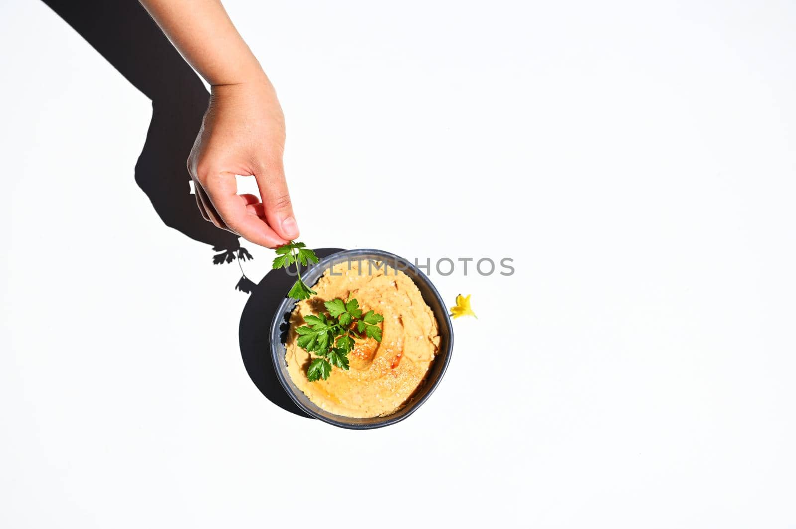 View from above of a hand holding parsley next to a bowl with hummus, isolated over white background with copy space for text
