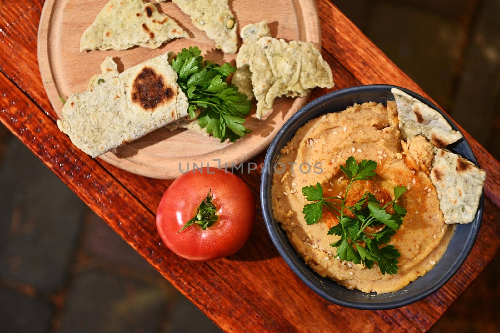 Overhead view of a bowl of a vegetarian chickpea hummus with sprinkled paprika and olive oil ripe red tomato and a wooden board with parsley wrapped in pit bread, on a rustic wooden table background