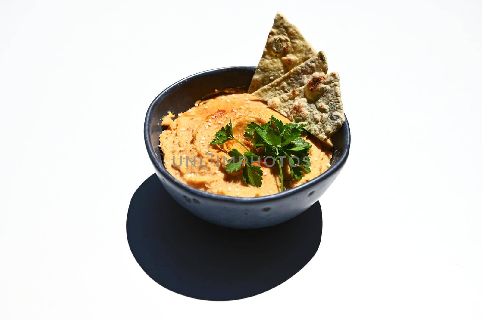 Close-up of a blue ceramic bowl with chickpea hummus, sprinkled with red chili and parsley with pita bread, isolated on white background with copy space for advertising text. Still life