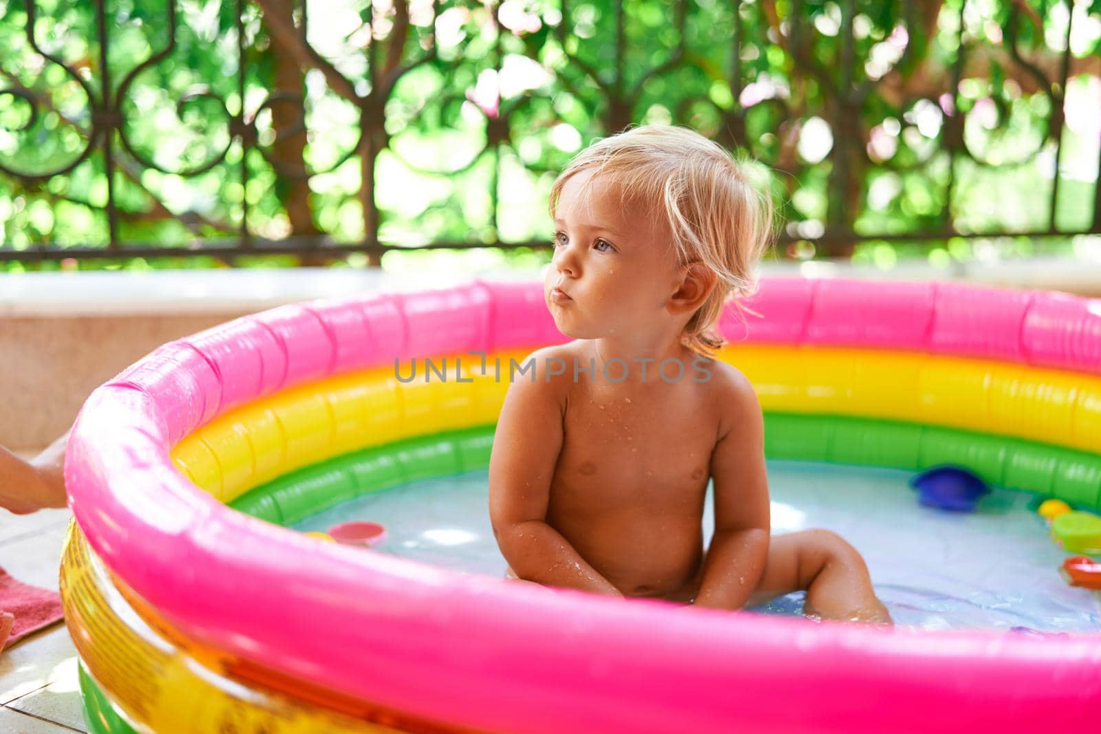 Little girl sits in an inflatable pool on the balcony with her head turned to the side by Nadtochiy