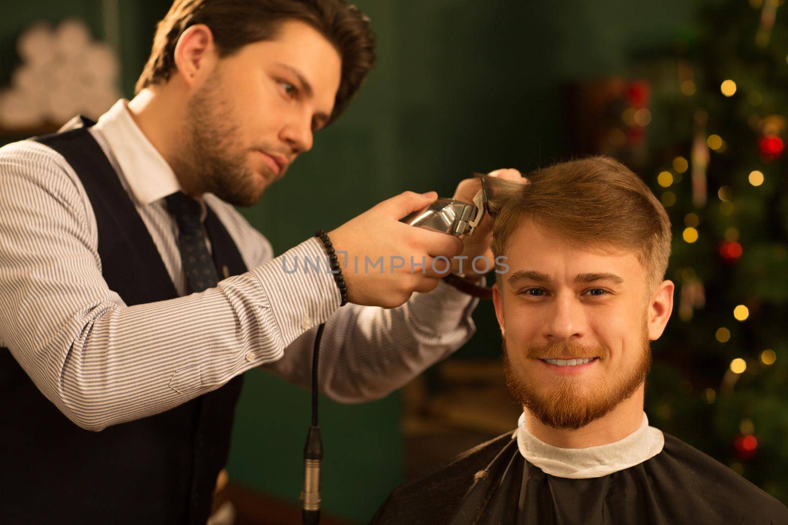Cheerful handsome young bearded man smiling joyfully to the camera while getting a new haircut by professional barber at the local barbershop copyspace customer client service lifestyle wellbeing grooming.