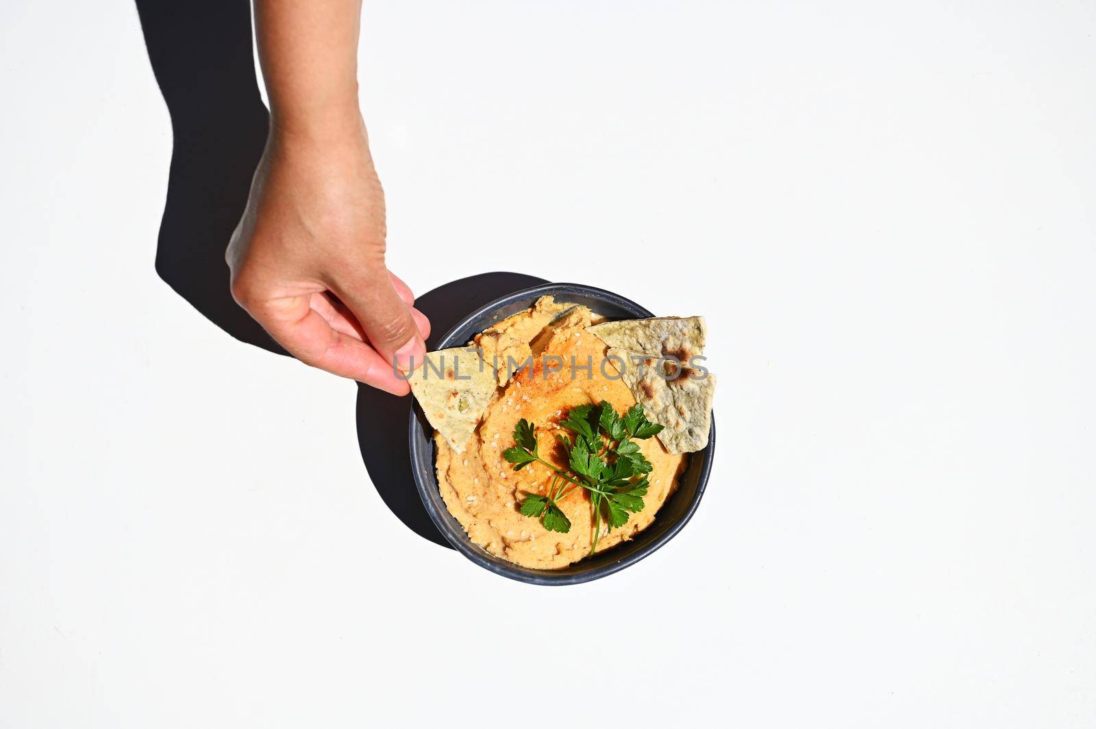 Overhead view of a hand, dipping pita bread into a creamy consistency vegan dish - oriental hummus with parsley in blue ceramic bowl, isolated on white background