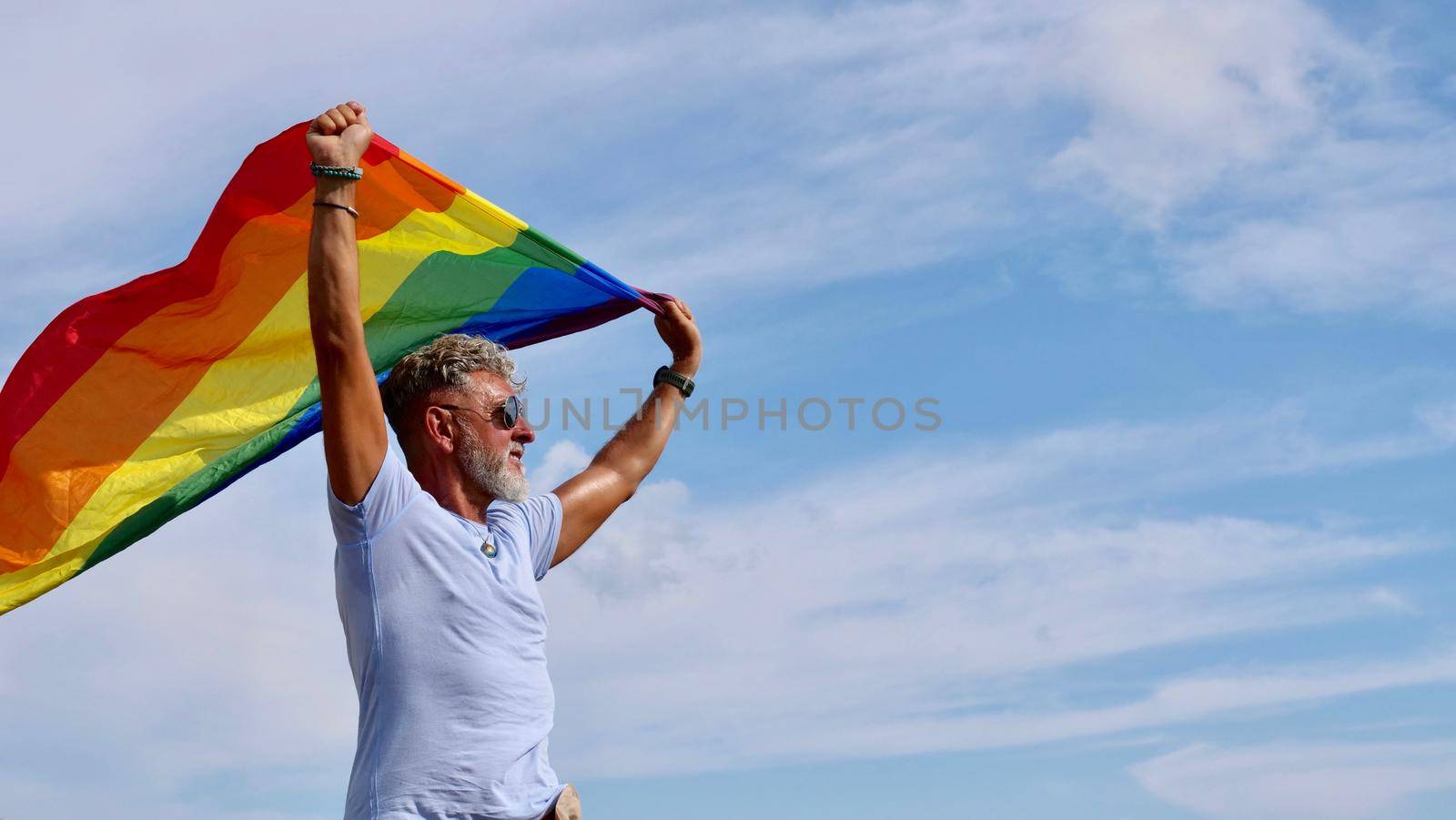 Portrait senior of a gray-haired elderly Caucasian man with a beard and sunglasses holding a rainbow LGBTQIA flag against a sky background in gay parade. Celebrates Pride Month, Coming Out Day