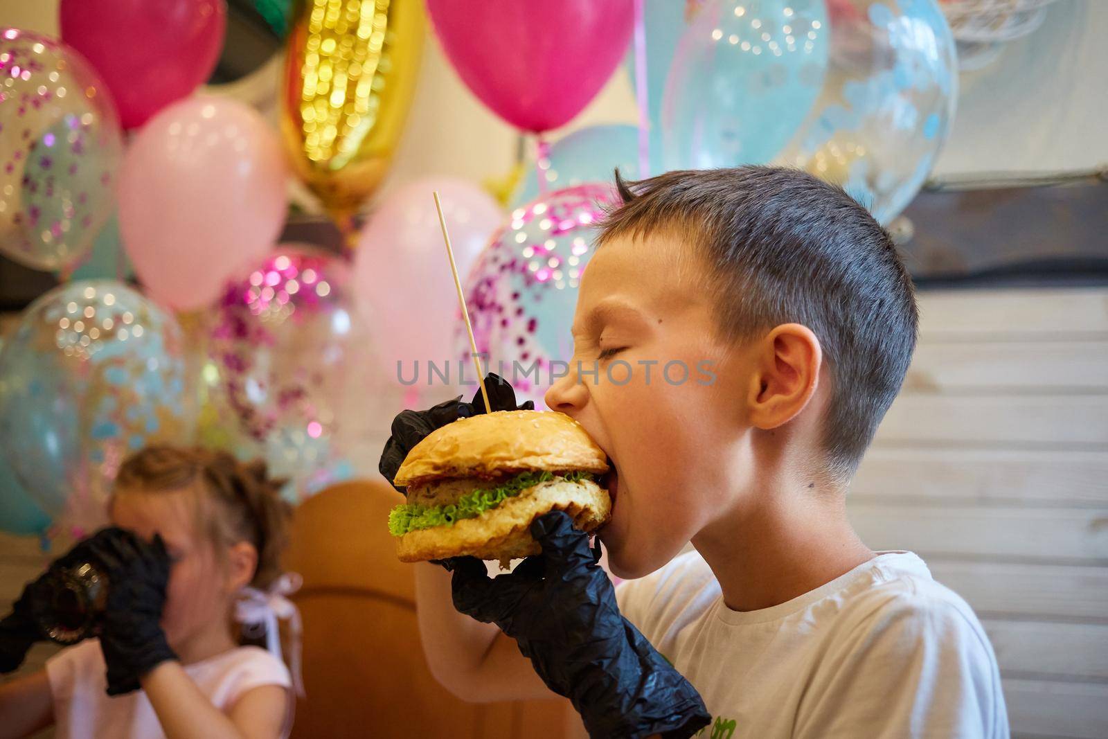 The handsome little boy eating burger in black rubber gloves