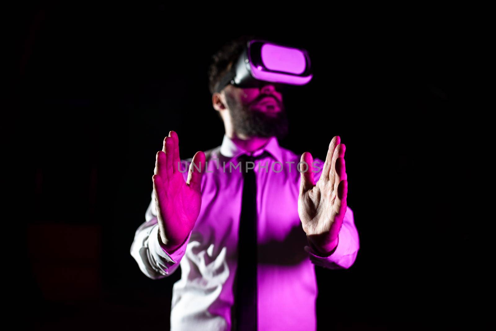 Bearded Businessman Wearing Virtual Reality Goggles And Enjoying Simulator. Light Falling On Man Using Futuristic Gadget Gesturing And Presenting Modern Innovative Technology. by nialowwa