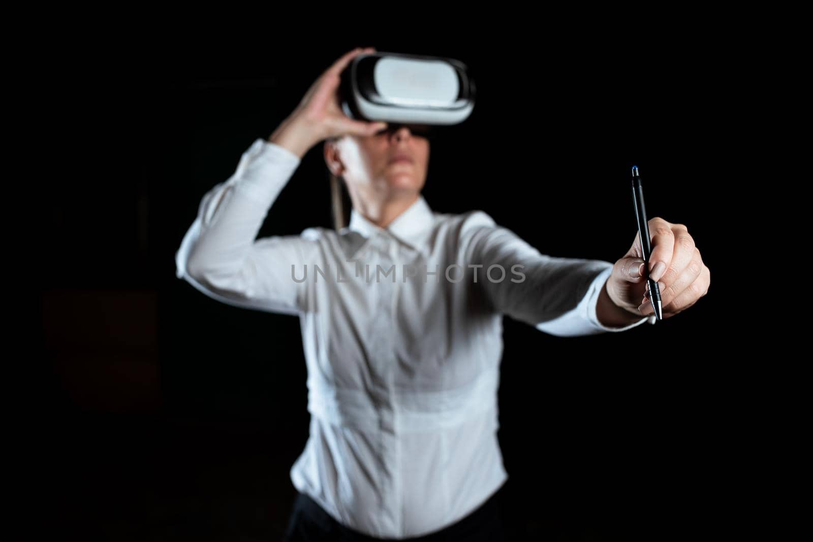 Businesswoman Wearing Virtual Reality Simulator And Holding Pen. Woman Wearing Vr Headset While Attending Professional Training Through Modern Technology. by nialowwa