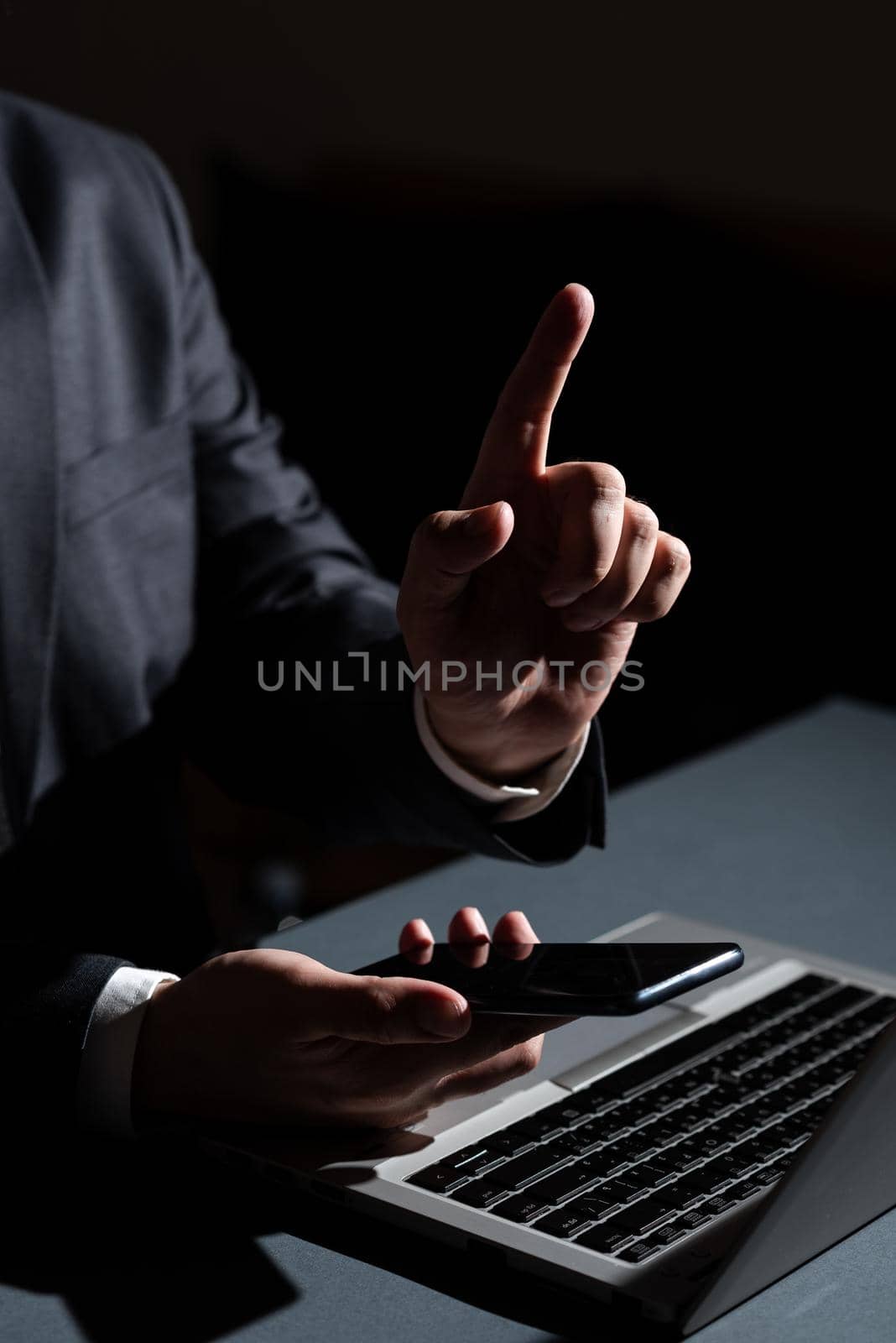 Businessman Holding Mobile Phone In Hand And Pointing With One Finger On Important Message On Desk With Lap Top. Man Having Cellphone And Presenting New Ideas With One Hand. by nialowwa