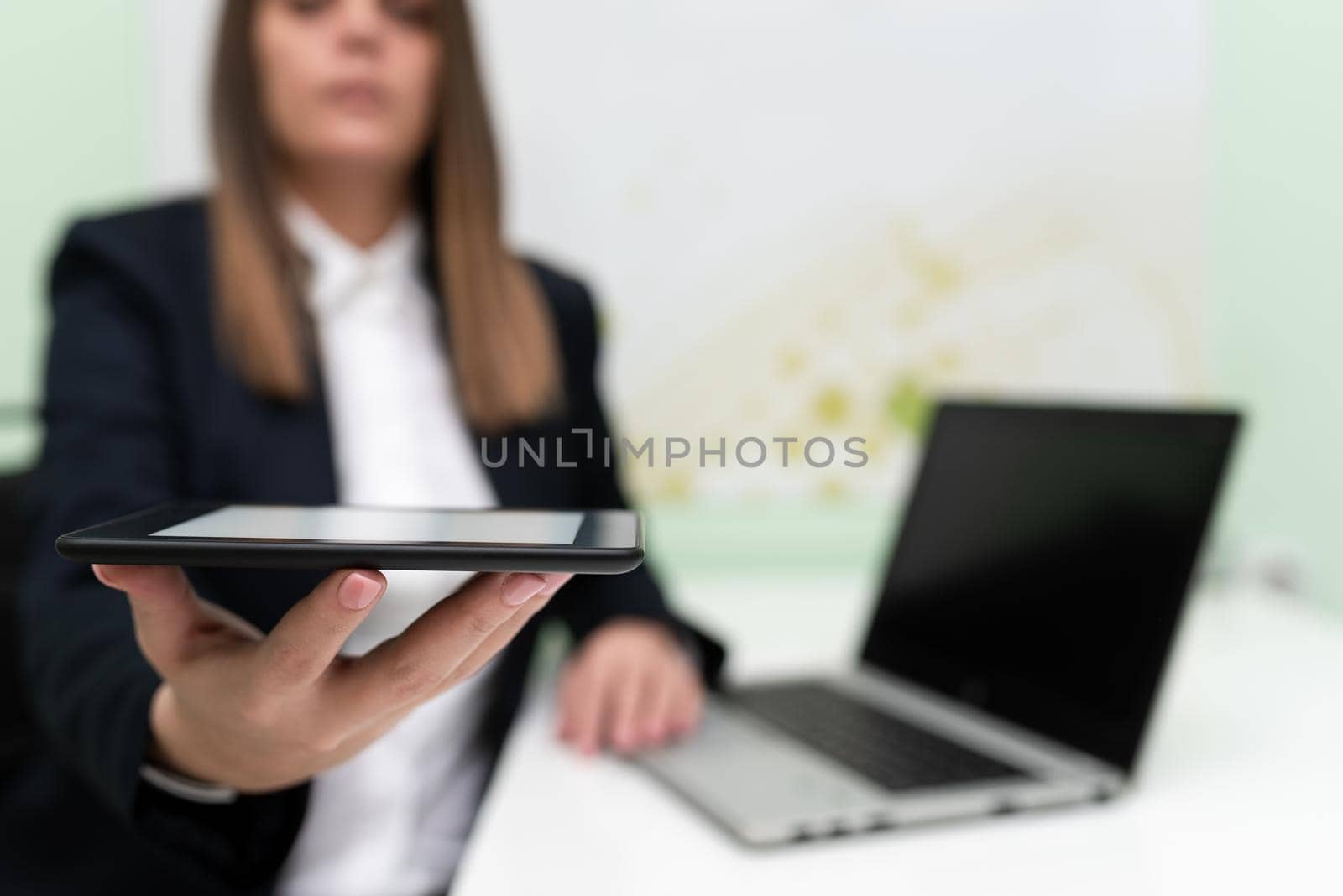 Businesswoman Holding Tablet With One Hand And Having Lap Top On Desk. Woman Presenting Important Data On Phone Screen. Sitting Executive On Desk Showing New Ideas. by nialowwa