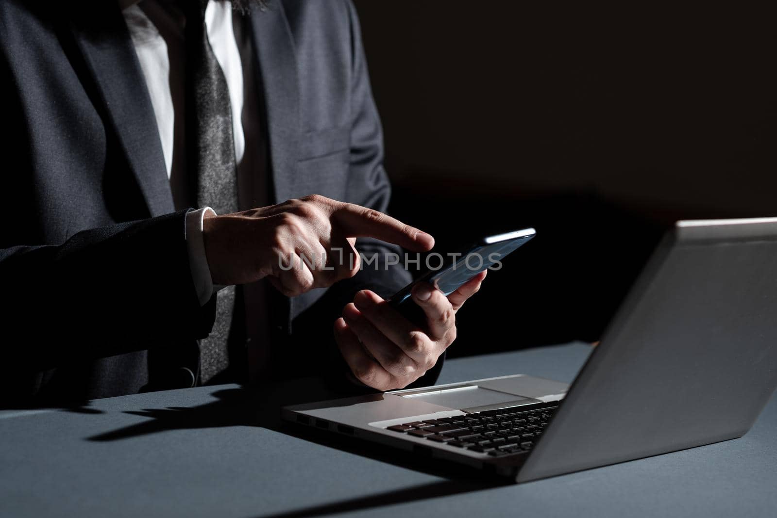 Businessman Holding Mobile Phone In Hand And Pointing With One Finger On It On Desk With Lap Top. Man Having Cellphone And Presenting Crutial Informations. by nialowwa