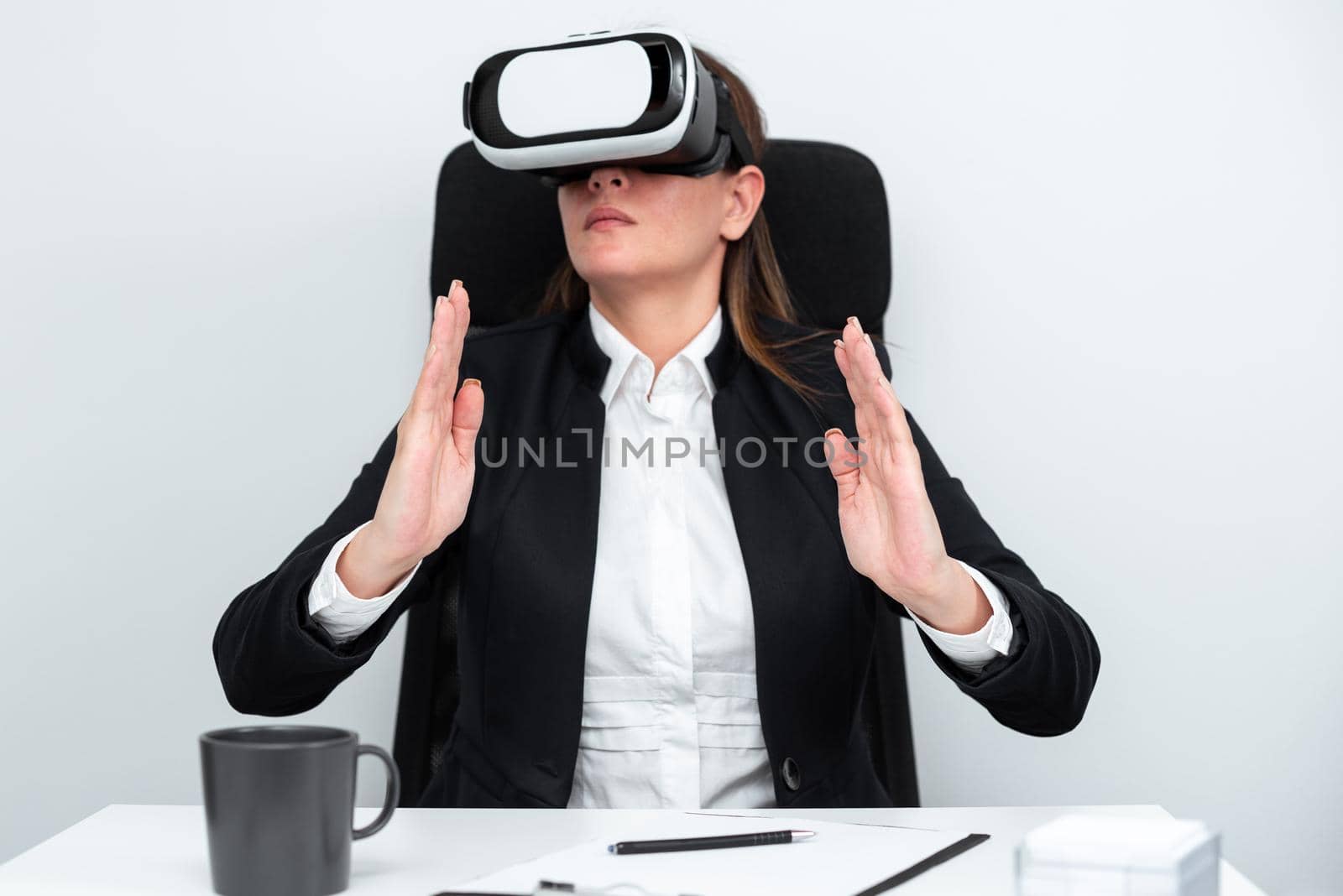 Female Executive Gesturing While Learning Professional Skill Through Virtual Reality Simulator. Woman Wearing Suit Sitting At Desk And Experiencing Modern Technology. by nialowwa