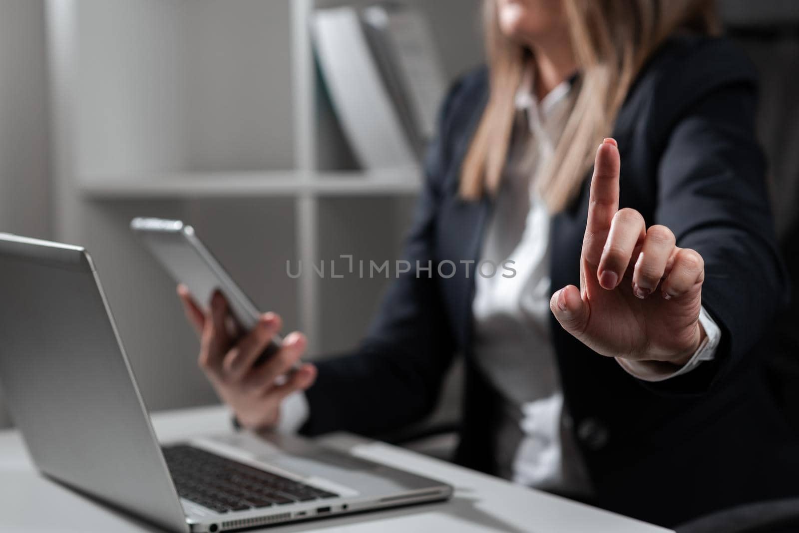 Businesswoman Holding Phone And Pointing With One Finger On Important Message On Desk With Lap Top. Executive In Suit Presenting Crutial Informations. by nialowwa