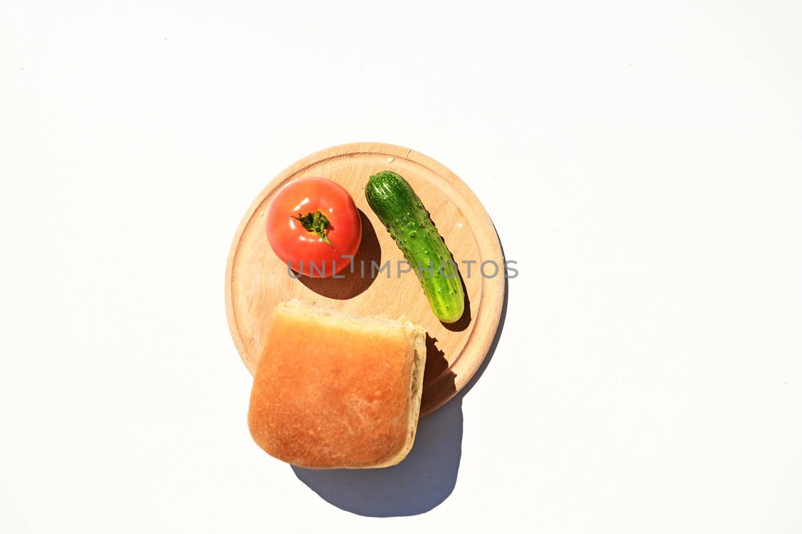 Top view of a wooden board with a loaf of freshly baked bread and raw tomato and cucumber, isolated over white background with copy space for advertising text. Food composition, still life. Flat lay