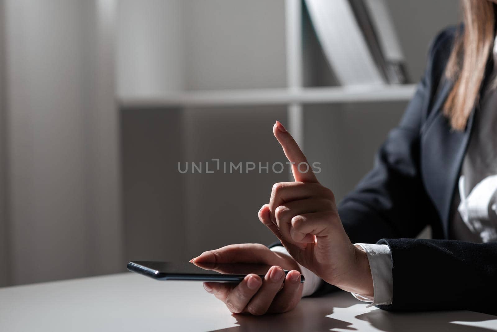 Businesswoman Holding Tablet In One Hand And Pointing With One Finger On Important Message. Sitting Woman In Office With Cellphone In Hand Presenting Informations. by nialowwa