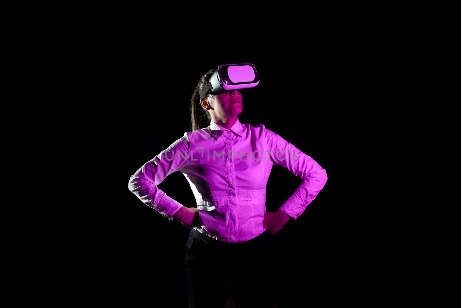 Female Manager Wearing Virtual Reality Headset And Taking Professional Training Through Modern Technology. Light Falling On Woman With Arms Akimbo Using Futuristic Gadget. by nialowwa