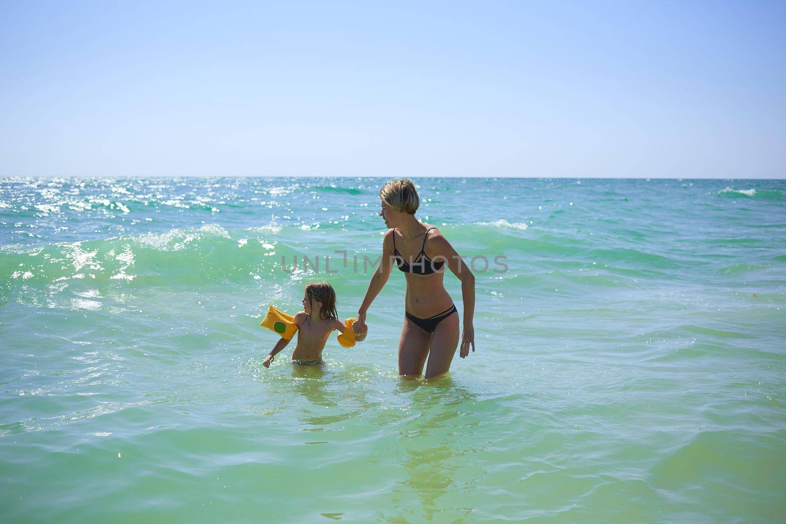Summer happy family of six years blonde child playing and jumping water waves embracing woman mother in sea shore beach.
