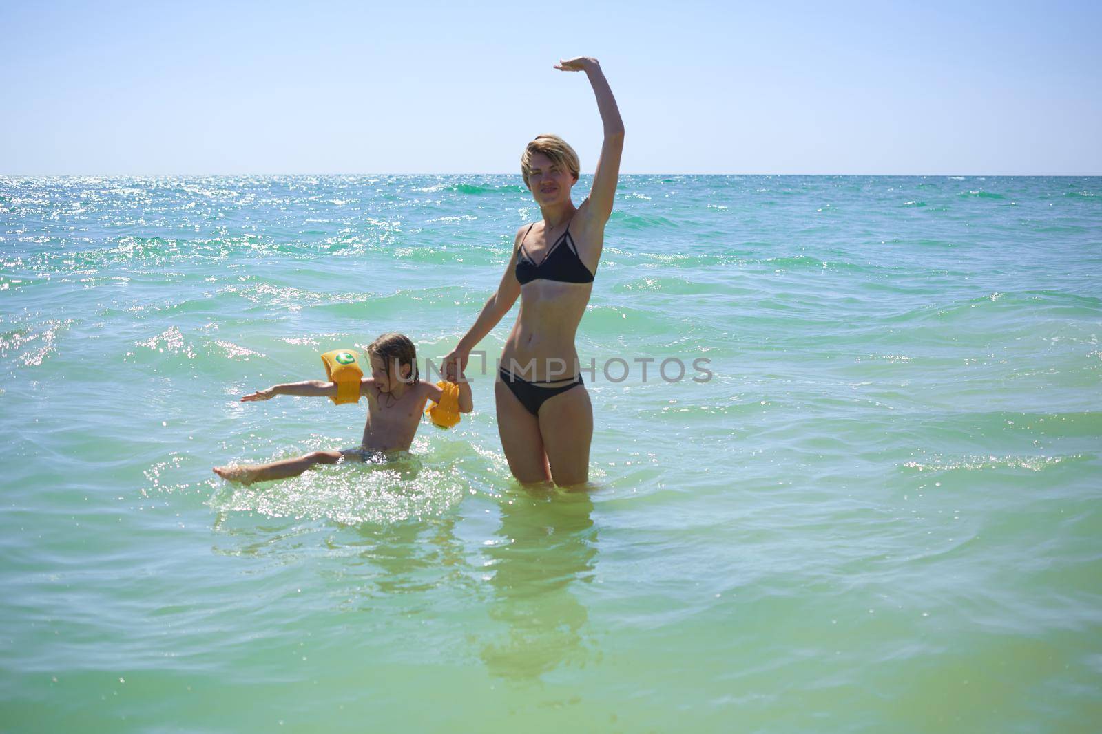 Summer happy family of six years blonde child playing and jumping water waves embracing woman mother in sea shore beach by sarymsakov
