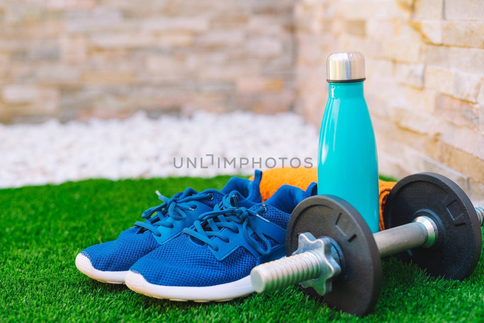 sports equipment in the foreground on green grass in the garden. orange towel, water bottle, weights and blue running shoes. stone wall background. by CatPhotography