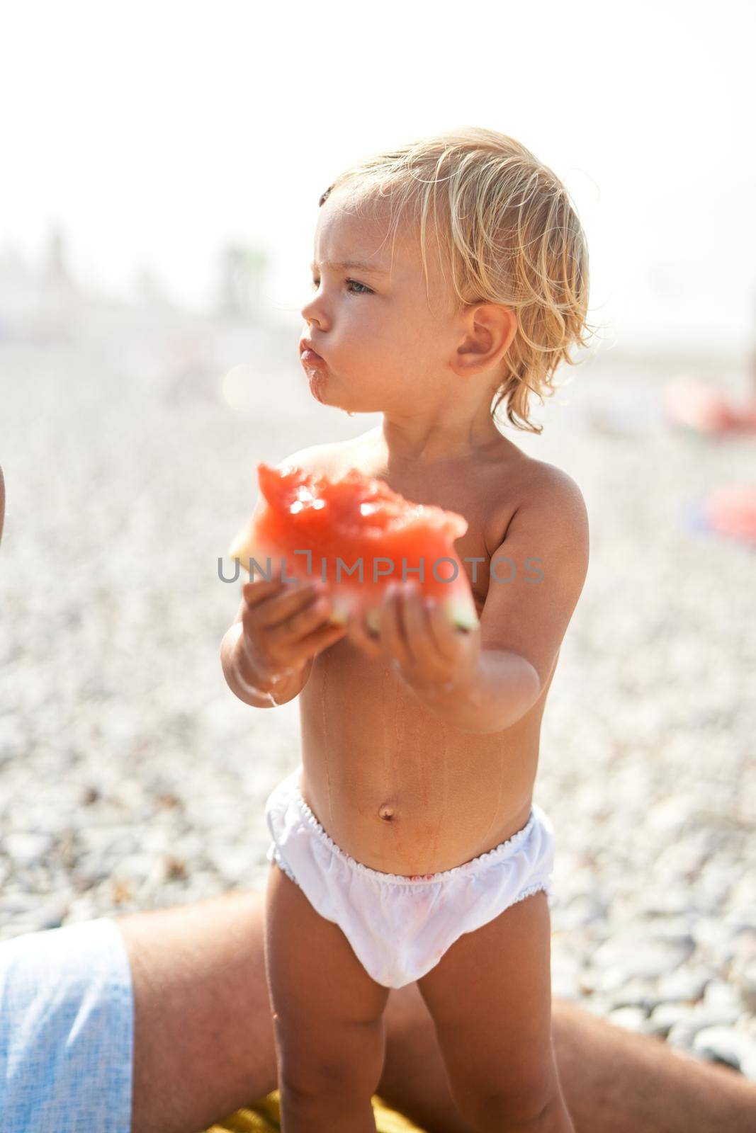 Little girl with a piece of watermelon stands on the beach and looks away by Nadtochiy