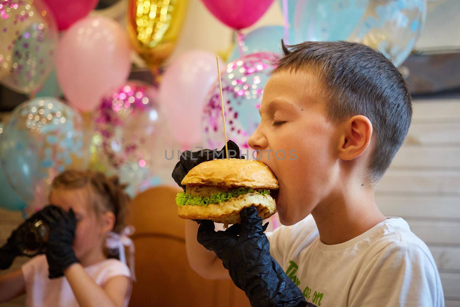 The handsome little boy eating burger in black rubber gloves