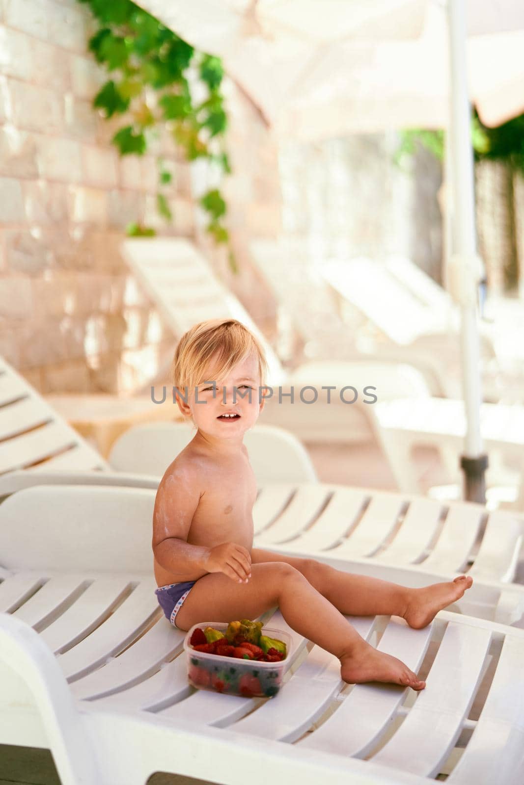 Little girl sits on a sun lounger in the shade near a fruit box by Nadtochiy