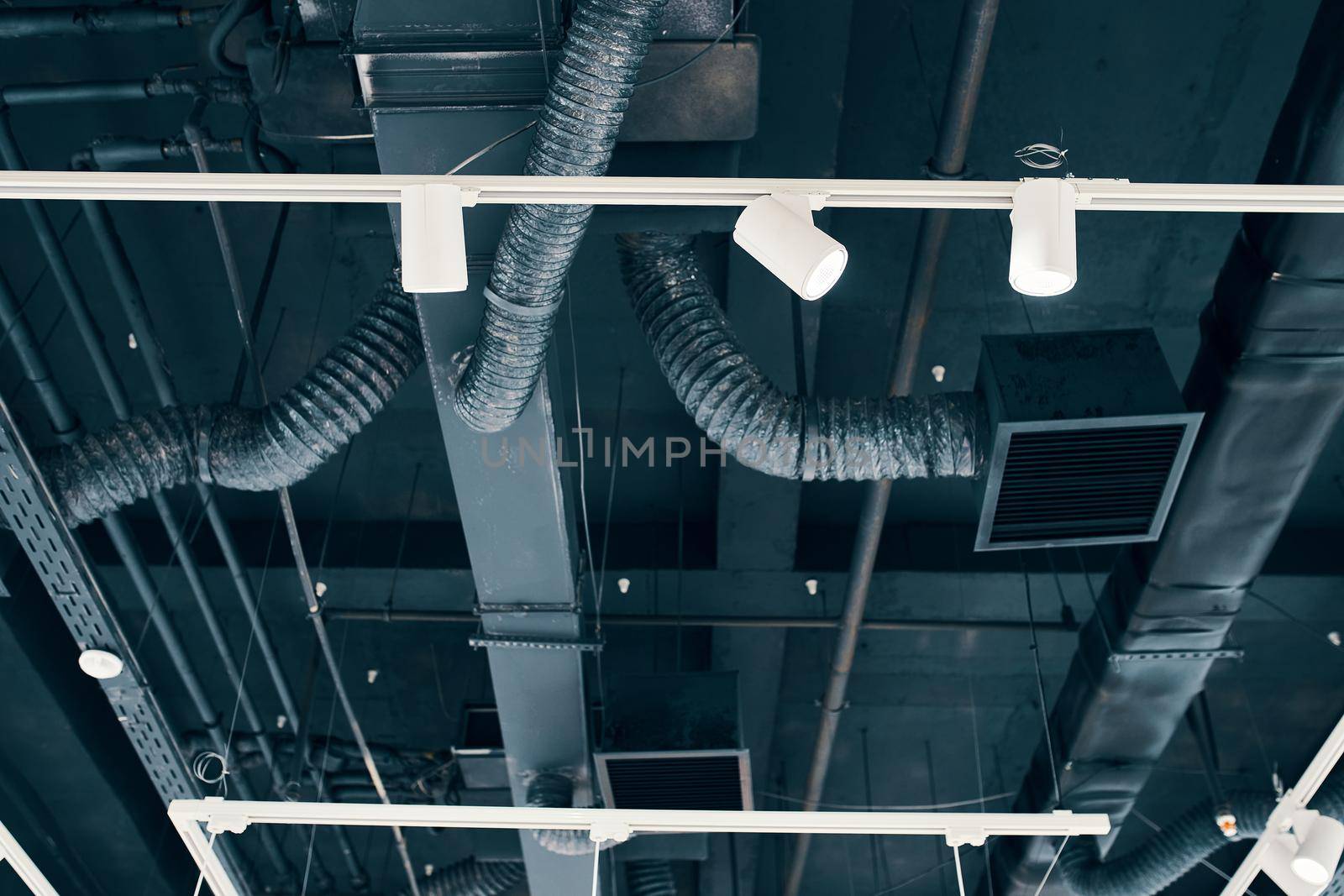 Ceiling mounted air condition units with other parts of ventilation system. Aluminum tubes, cables and vents located inside commercial hall with hanging lights and other construction parts.