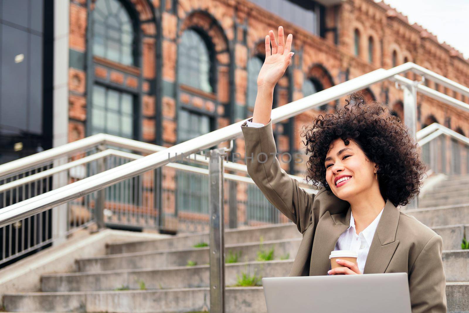 happy young woman greets smiling sitting in the city with a laptop and a coffee, concept of business and urban lifestyle, copy space for text