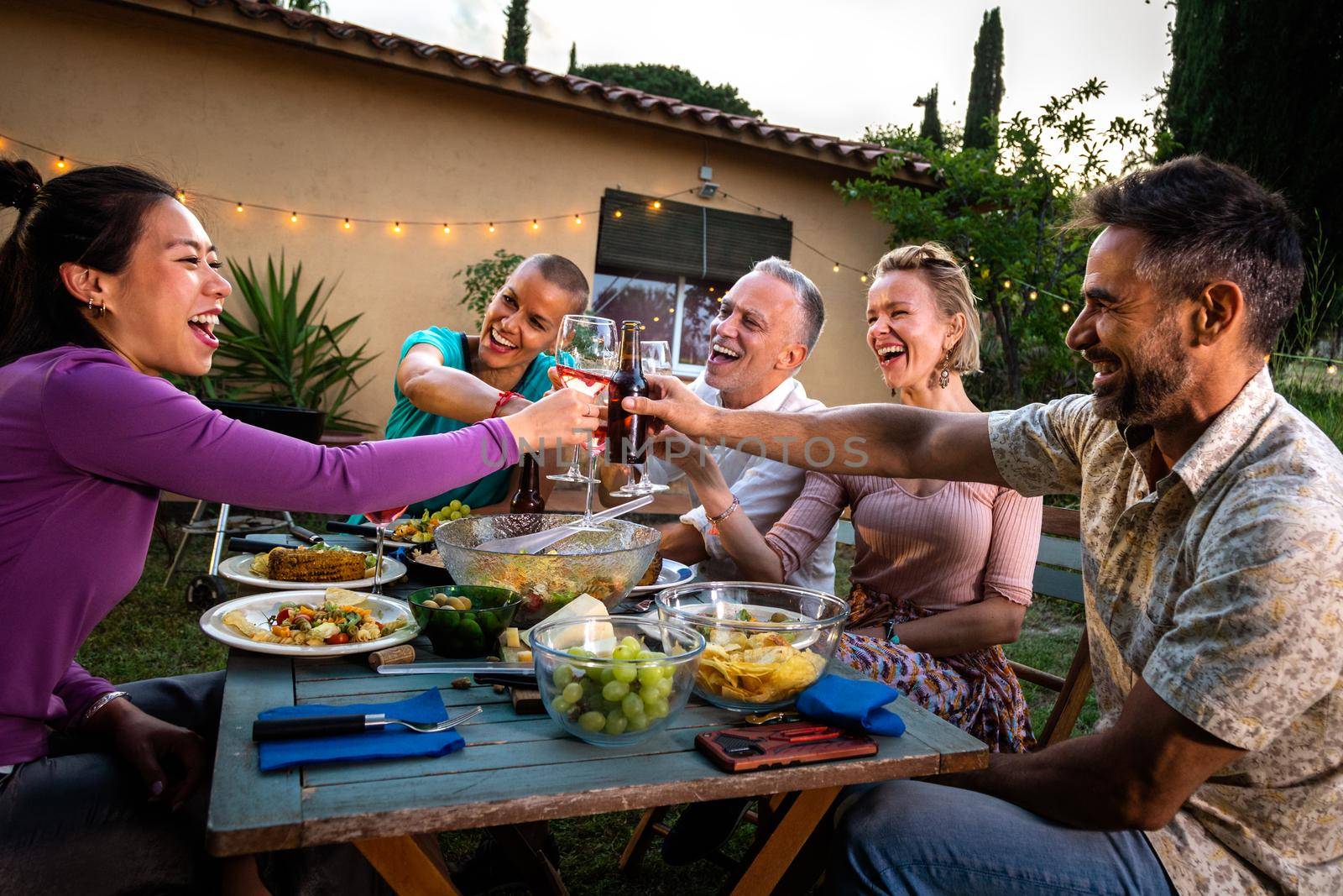 Asian woman having a celebratory toast with wine, laughing and having fun during barbecue garden dinner party at sunset. Friends having fun. Lifestyle.