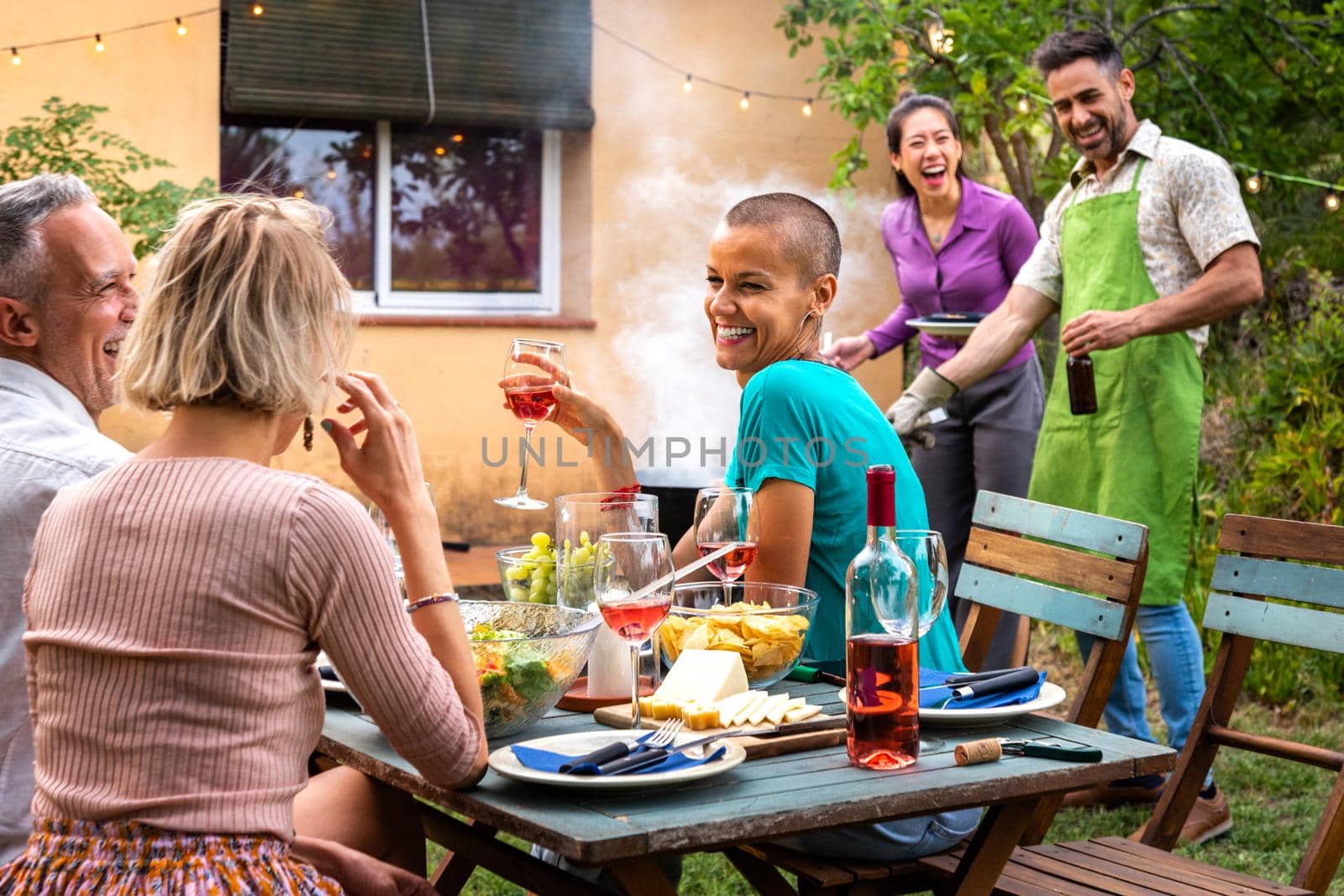 Multiracial couple cooking food on grill for friends. Outdoor garden barbecue party. Friends laughing and having fun. by Hoverstock