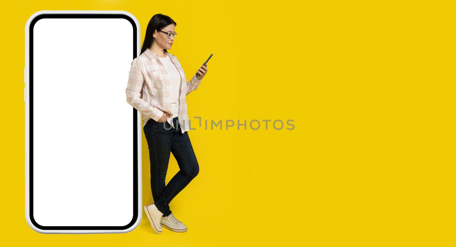 Leaned on big smartphone with white screen charming middle aged asian woman looking at phone in her hand wearing casual isolated on yellow background. Copy space on right, phone mock up.