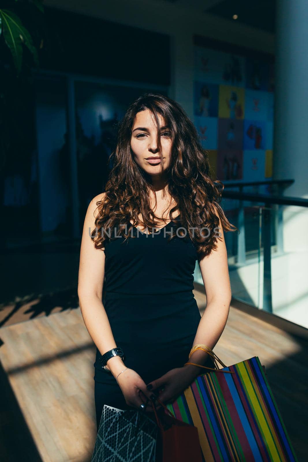 Portrait of a beautiful young woman in a black dress and curly hair in the mall who is shopping in shopping bags, looking at the camera, smiling