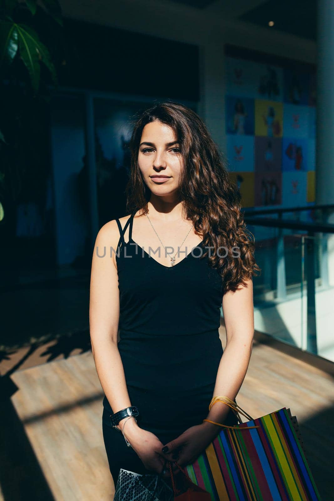 Portrait of a beautiful young woman in a black dress and curly hair in the mall who is shopping in shopping bags, looking at the camera, smiling