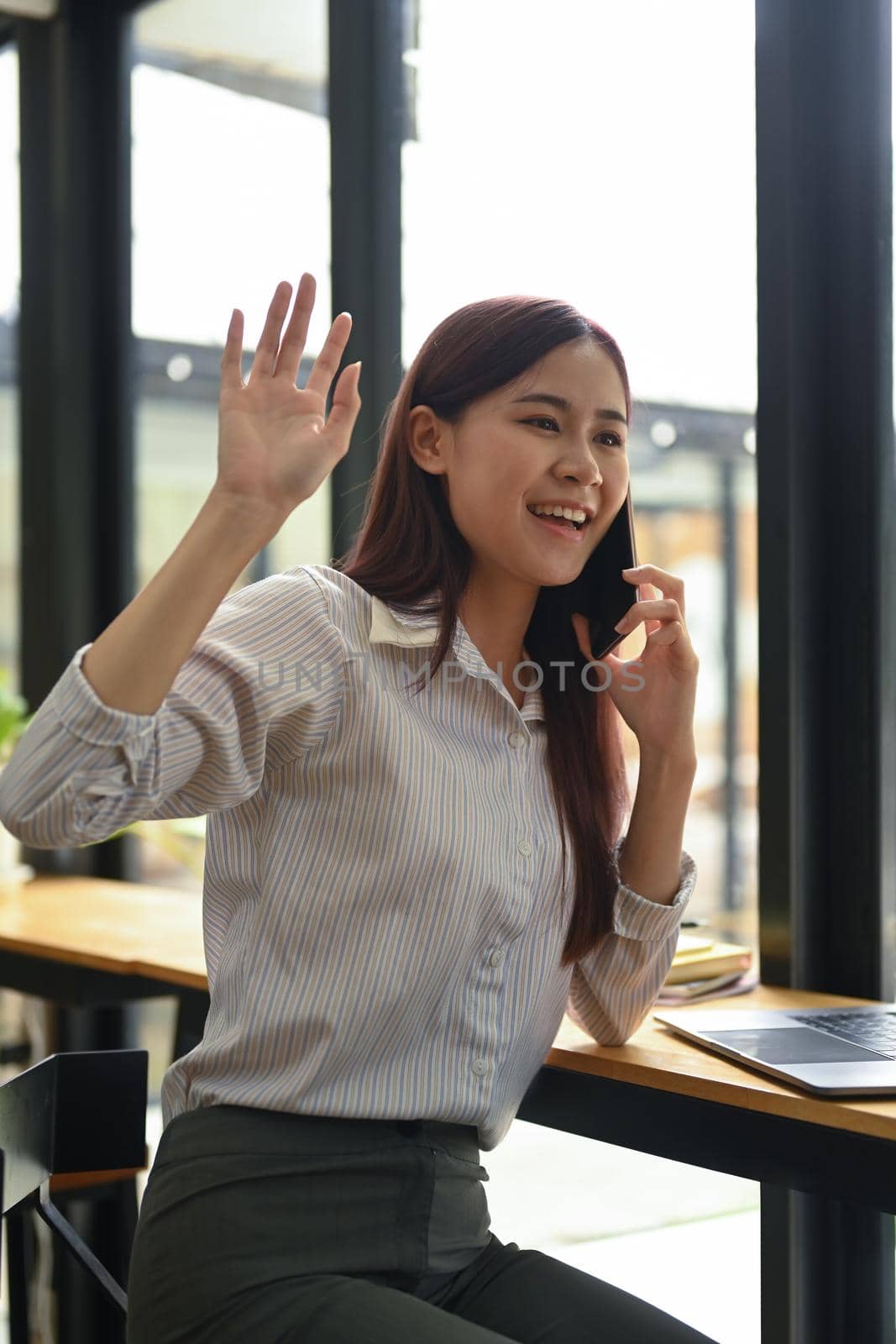 Cheerful female employee waving hands to greet her colleagues while talking on mobile phone by prathanchorruangsak