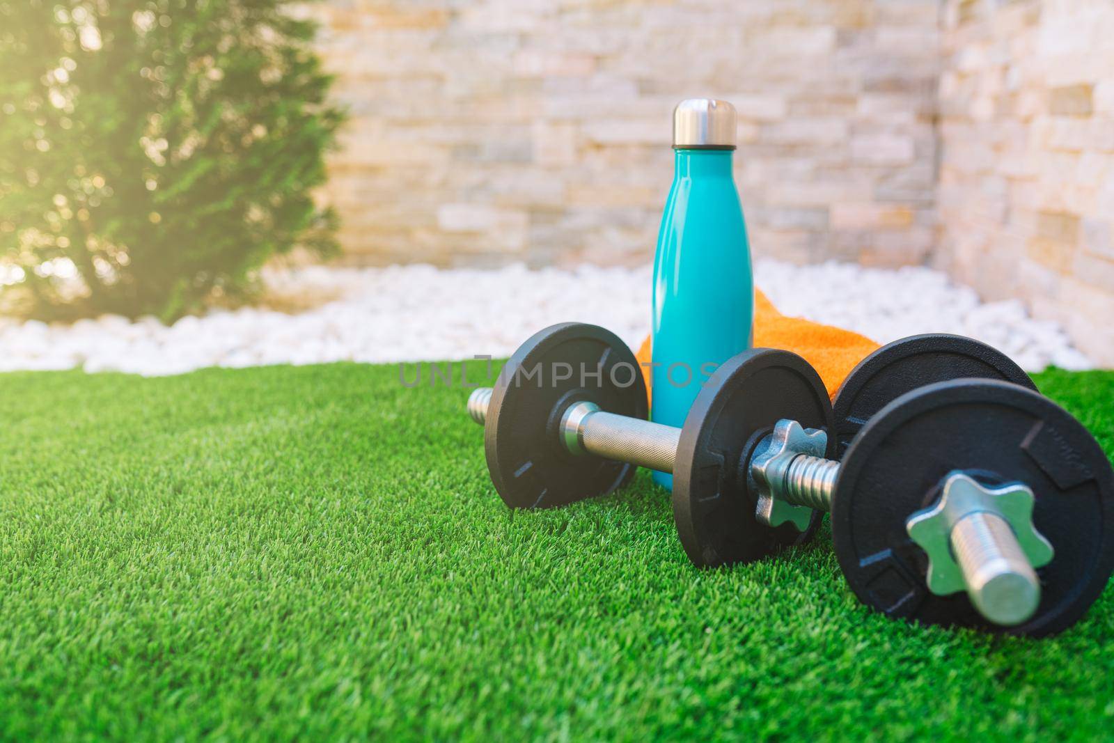 sports equipment on the green grass in the sunlit garden. orange towel, water bottle, weights. stone wall background. natural outdoor light.