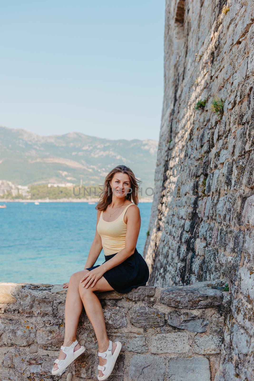 Beautiful girl sitting on a stone wall, in background is the blue sea, Budva, Montenegro