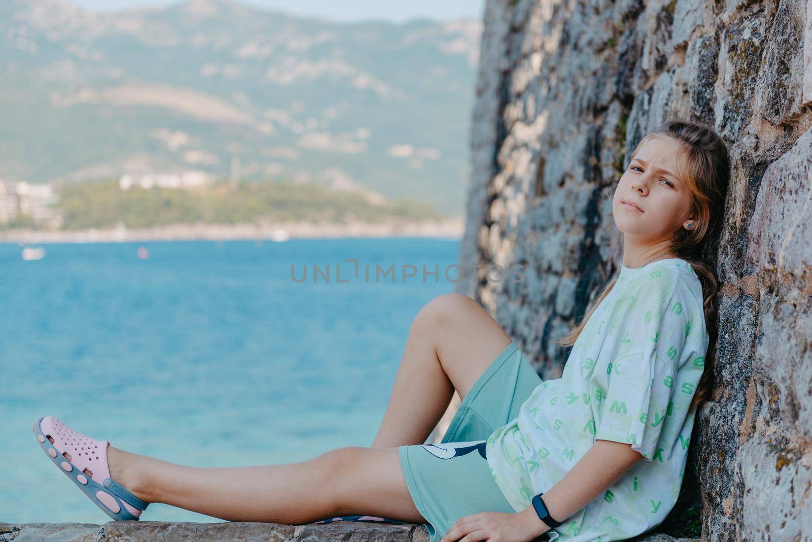 Beautiful girl sitting on a stone wall, in background is the blue sea, Budva, Montenegro. by Andrii_Ko