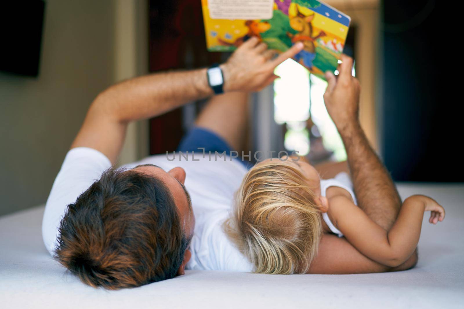 Dad shows his daughter a book with pictures lying on the bed. High quality photo