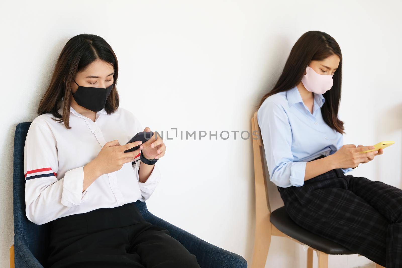 social distancing concept, Two women wearing masks and distancing while sitting on mobile phones following coronavirus social trend. by Manastrong