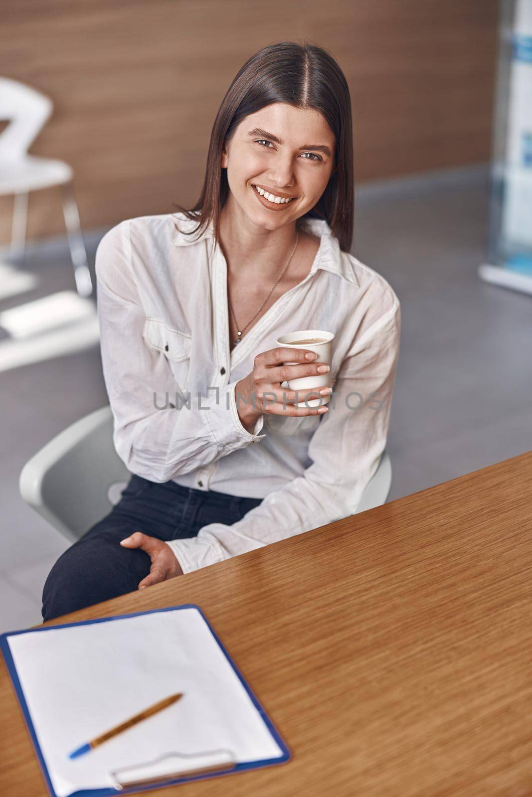 Joyful caucasian lady is drinking a coffee from a white paper cup while sitting on reception by Yaroslav_astakhov