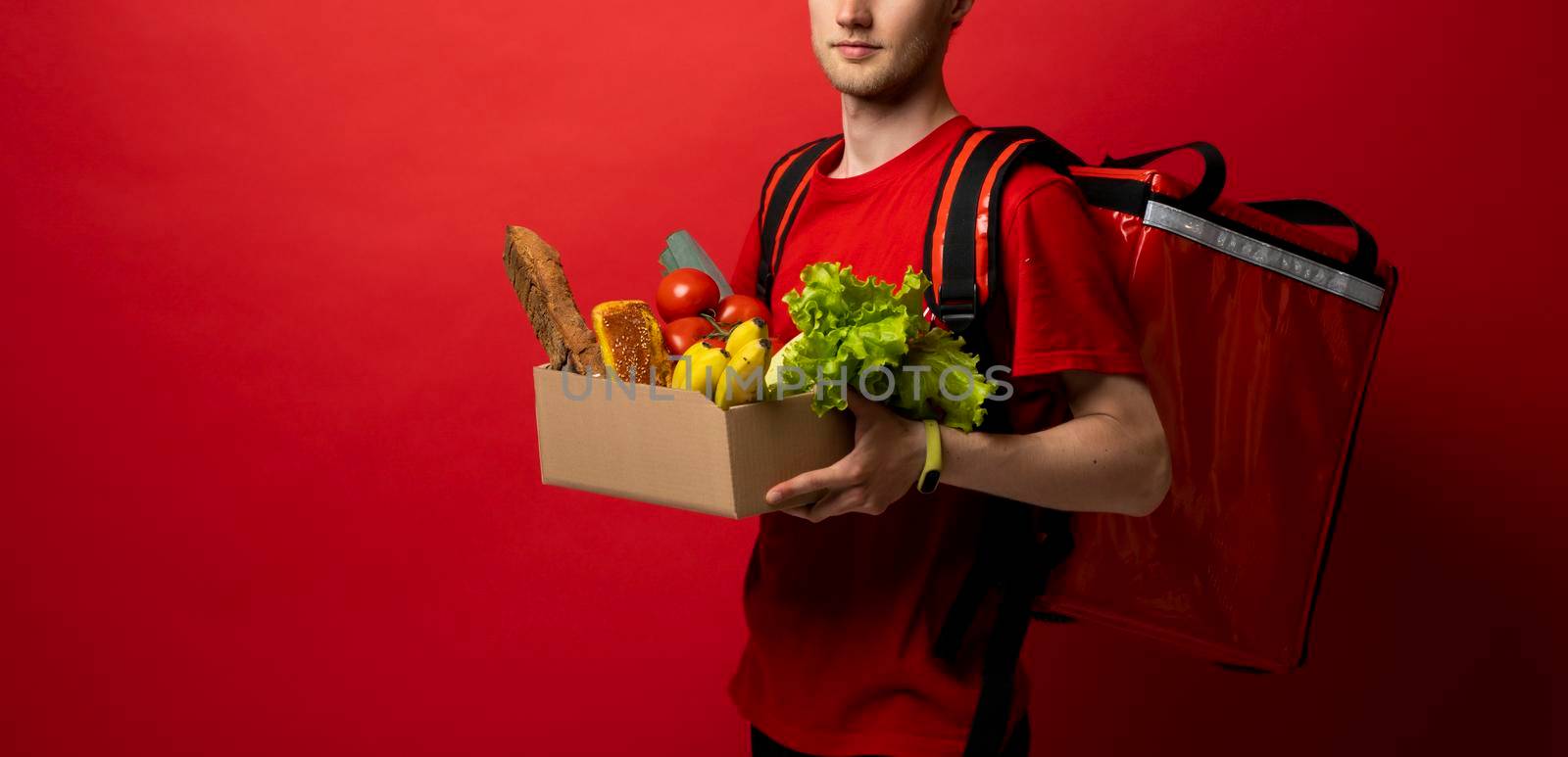 Crop picture of delivery man in red uniform carrying paper box with food products isolated over red background. Food delivery service