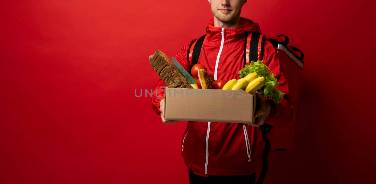 Male delivery man in red uniform holds paper box with a vegetables on red background. by vovsht