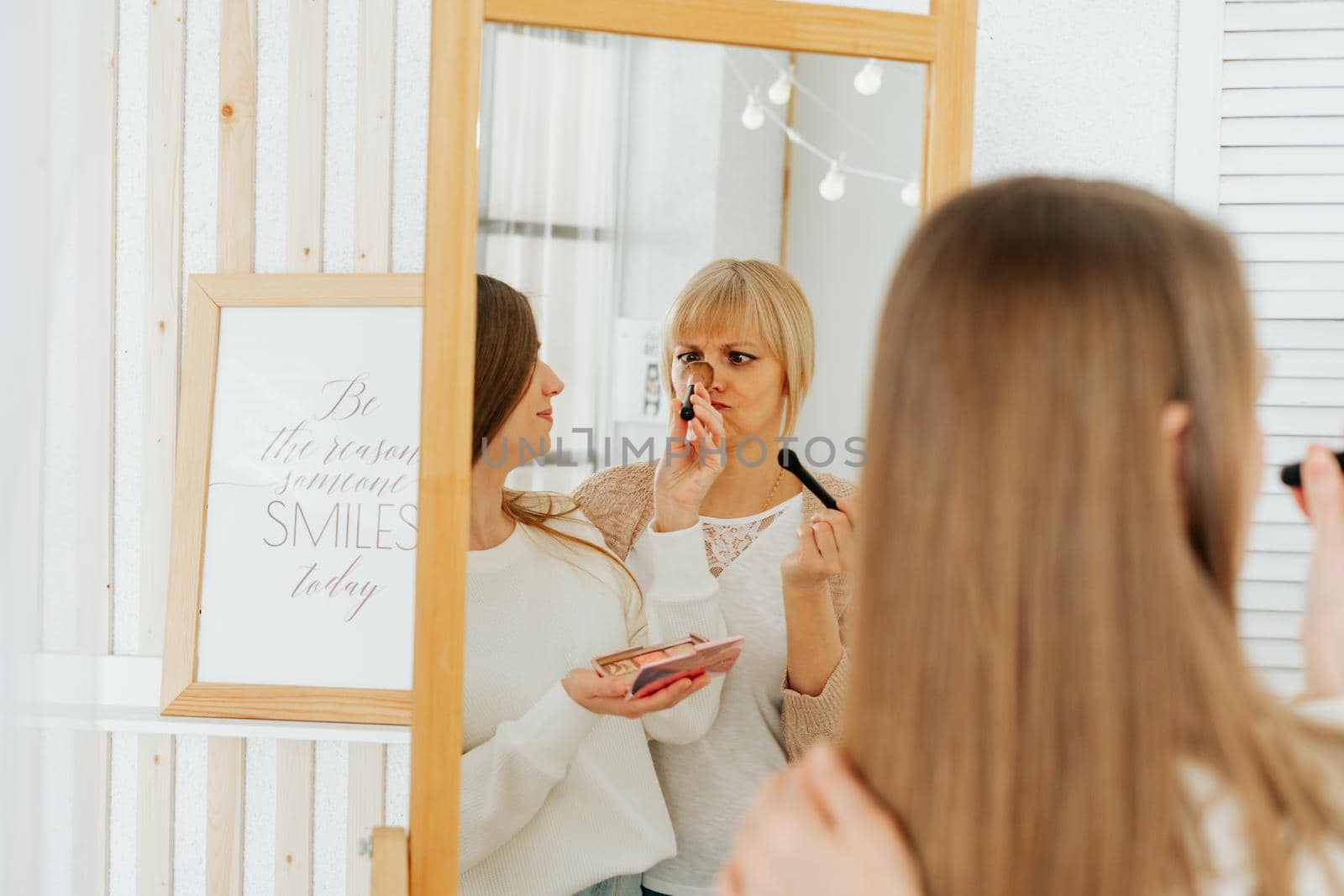 Beautiful smiling young caucasian mature senior mother mom and adult daughter doing makeup, standing near mirror, and looking at reflection in living room home. Family with two women relax together.