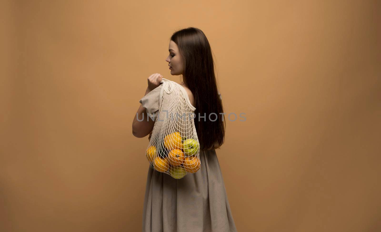 Back of woman in beige dress holding string shopping reusable mesh bag with fruits on a shoulder. Zero waste concept. Space for text. Reusable eco friendly object. Copy space