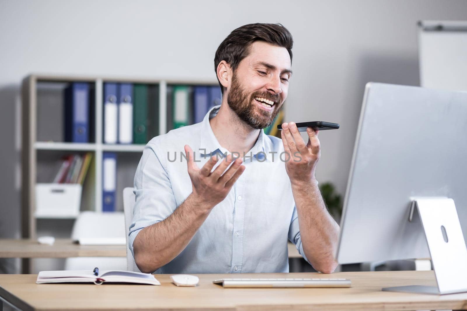 Young man with a beard, freelancer, manager, worker talking on a cell phone in the office, sitting at a computer desk, talking, waving